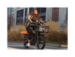 A woman wearing a black outfit with green jacket and a black bike helmet riding a green electric bike on a road in a city setting. 