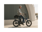 A man with tattoos wearing a plaid shirt, jeans, and a grey helmet smiling and sitting on an ebike in an urban setting. 