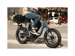 A man riding a blue electric bicycle with an orange Rad Power Bikes logo, fat black tires, and large basket with a cargo net, on a paved road in an urban setting. 